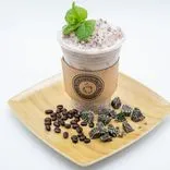 Iced coffee in a plastic cup with a mint leaf garnish, on a wooden tray with scattered coffee beans and chocolate candies.