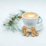 A cup of cappuccino with heart-shaped latte art accompanied by two gingerbread cookies and pine branches with festive decorations on a white background.