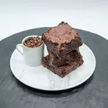 A stack of three chocolate brownies on a white marble plate with a small white cup filled with coffee beans, on a black surface with a white background.