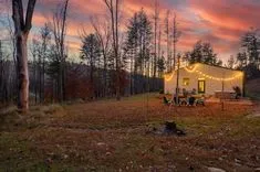 Rustic cabin at twilight with string lights, surrounded by autumnal trees and Adirondack chairs around a fire pit.