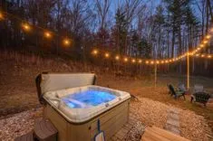 Outdoor hot tub with illuminated water, surrounded by string lights in a wooded backyard at dusk.