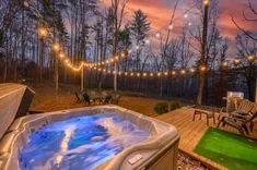 Cozy outdoor deck with a hot tub and string lights at dusk, surrounded by bare trees and a BBQ grill to the side.