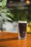 A cold glass of dark beer with foam on top, sitting on a wooden table with blurred green plants in the background.