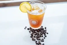 Iced coffee beverage garnished with a lemon slice, surrounded by scattered coffee beans on a light surface.