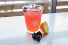 Iced berry lemonade in a glass with a logo, blackberries, and a lemon slice on a white table with a blurred background.