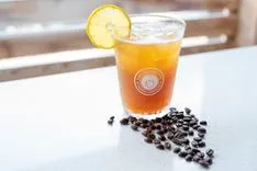 Iced coffee in a branded glass garnished with a lemon slice, surrounded by coffee beans on a table.