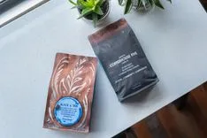 Two bags of coffee on a kitchen counter next to potted plants, one with a brown design labeled 'Black & Tan' and the other black labeled 'COFFEEHOUSE FIVE'.