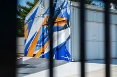 Colorful geometric mural on shipping containers viewed through a fence.