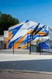 A vibrant blue, orange, and white geometric mural painted on the side of an industrial building behind a black metal fence, with clear blue sky in the background.