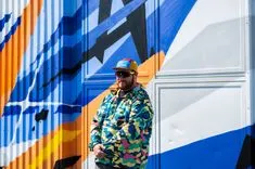 Man in a colorful camouflage jacket and cap standing in front of a vibrant, geometric mural.