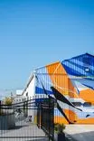 Colorful geometric mural painted on the exterior of an industrial building behind a black metal fence, with a clear blue sky above.
