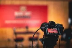 Digital camera on a tripod with a blurred background showing a red banner with text.
