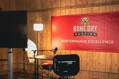Podcast recording setup with microphone and headphones in front of a "Bone Dry Roofing - Performance Excellence" banner.