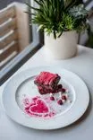 A gourmet dessert of a chocolate brownie dusted with red powder, garnished with white chocolate chips and raspberries on a white plate, with a potted succulent plant in the background next to a window.