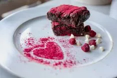A stack of chocolate brownies with raspberry dust and dried raspberries on a white plate with a heart-shaped dusting decoration.