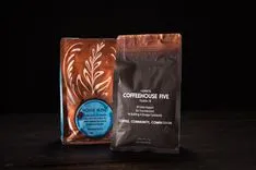 Two bags of coffee from Coffeehouse Five on a dark wooden surface, one labeled 'House Blend Medium Roast' with a decorative design, the other promoting community support and compassion.