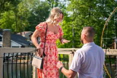 Woman in a floral dress surprised by kneeling man holding a small box outdoors.