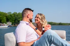 Couple sitting close together on a boat, smiling and gazing into each other's eyes, with a lake and trees in the background.
