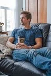 Man sitting on a leather couch holding a coffee cup, looking contemplatively out a window.