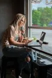 Woman sitting at a table by the window in a cafe, drinking coffee while working on a laptop.