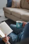 Person sitting on the floor holding a coffee mug and reading a book with a couch in the background.