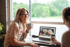 Two women enjoying a conversation over coffee with a laptop open on the table in a cafe setting.