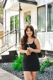 A smiling woman in a black dress holding an iced coffee standing in front of a house with a white facade and black trim.