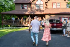 Two people walking towards a house where a group of guests are gathered on the porch for a social event.