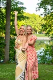 Two women in summer dresses smiling and posing with one raising a surfboard in a park with trees and a pond in the background.