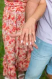 Close-up view of a couple holding hands, showcasing an engagement ring, with a floral dress and blue jeans in the background.