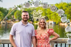 Couple standing together with a "Better Together" neon sign surrounded by white flowers, with a lake in the background.