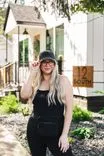 Woman in black outfit and hat standing in front of a house with lush greenery.