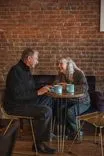 An older couple smiling at each other and holding mugs over a small round table in a cozy cafe with a brick wall in the background.