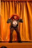 Person on stage with hands on head, wearing a curly red wig, looking surprised in front of a yellow curtain.