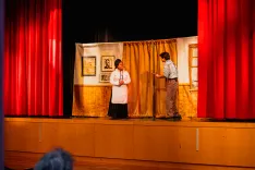 Two actors performing on a stage with a simple backdrop, simulating a room with red curtains on the sides.