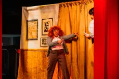 Two actors performing on stage, one with a curly wig pointing excitedly at another partially obscured by a curtain.
