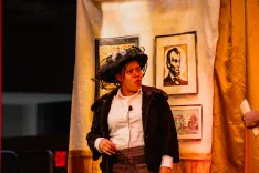 Actor on stage in period costume with hat, standing beside a painting, expressing surprise or confusion.