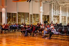 Audience seated on chairs watching a performance in a warmly lit indoor space with decorative lights.