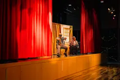 Two actors performing on a stage with a backdrop of a room, framed by red curtains.