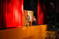 Two actors performing on stage with red curtains and stage lighting.