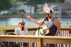 Two musicians performing on a wooden deck by a lake, one playing guitar and the other singing with a microphone.