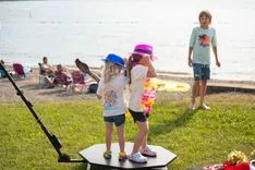 Two young children wearing colorful hats standing on a board by a lake while another child watches from the background.