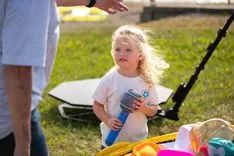 A toddler holding a water bottle looks up at an adult out of frame, with toys scattered on the ground nearby in a park setting.