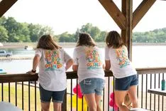 Three people standing on a covered porch overlooking a lake, wearing matching t-shirts and facing away from the camera.