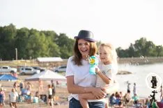 Woman in a hat smiling and holding a young child at a busy outdoor event, with cars and people in the background.