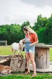 Woman in a casual outfit petting a goat on a log stump with another goat laying down beside them.