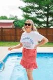Woman in white t-shirt and red checkered skirt standing by a swimming pool, wearing sunglasses and smiling.