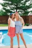 Two smiling young women standing by a poolside, one wearing a red checkered skirt and a white tank top with a blue headband, and the other in a blue gingham romper with a red headband.
