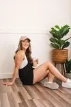 Young woman in a cap and casual outfit seated on a wooden floor with a potted plant in the background, holding an iced coffee and smiling.