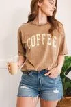 Woman in a brown T-shirt with the word 'COFFEE' holding an iced coffee while standing in a room with a plant in the background.